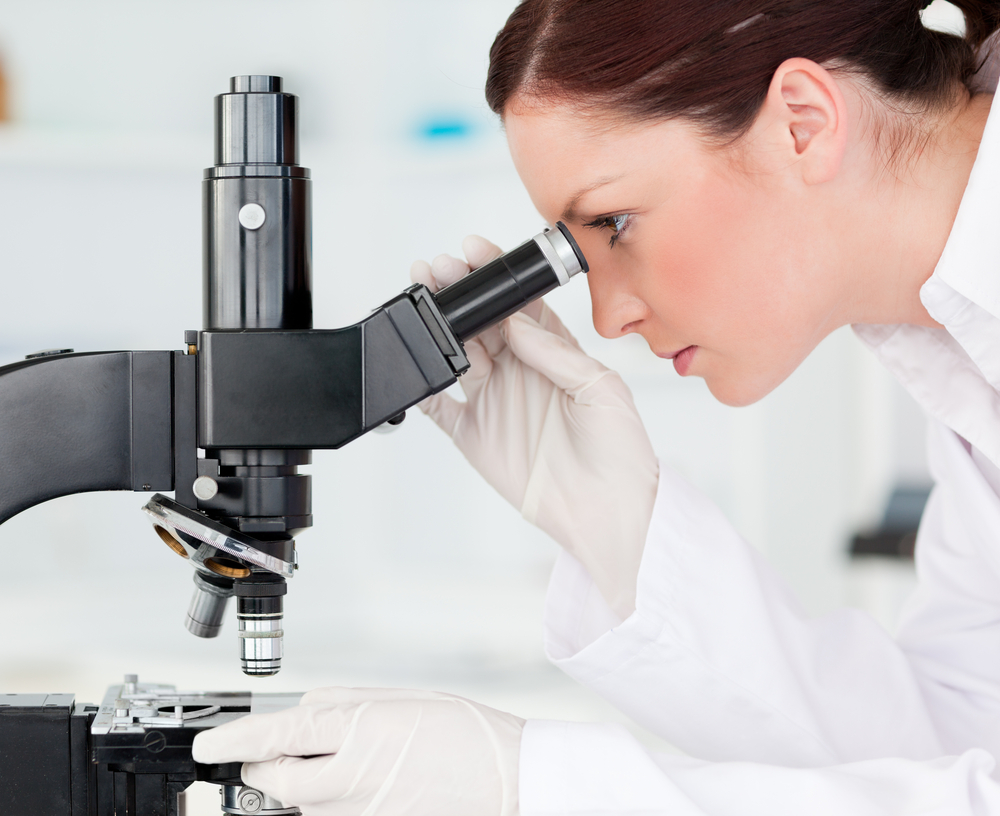 woman in white coat and gloves with hair tied back looking into a microscope
