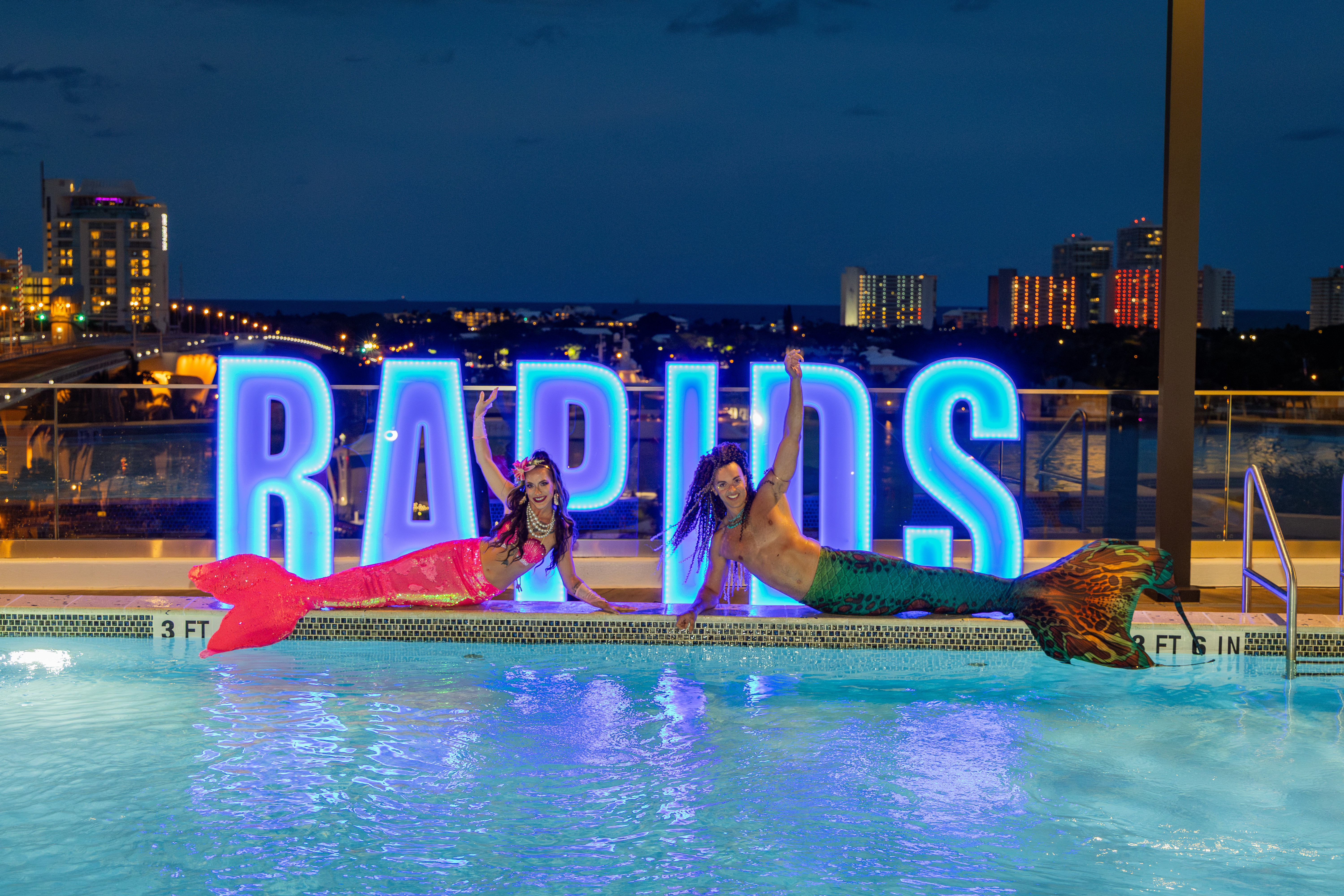 mermaid and merman with lighted rapids sign on omni ft lauderdale pool deck with night time skyline