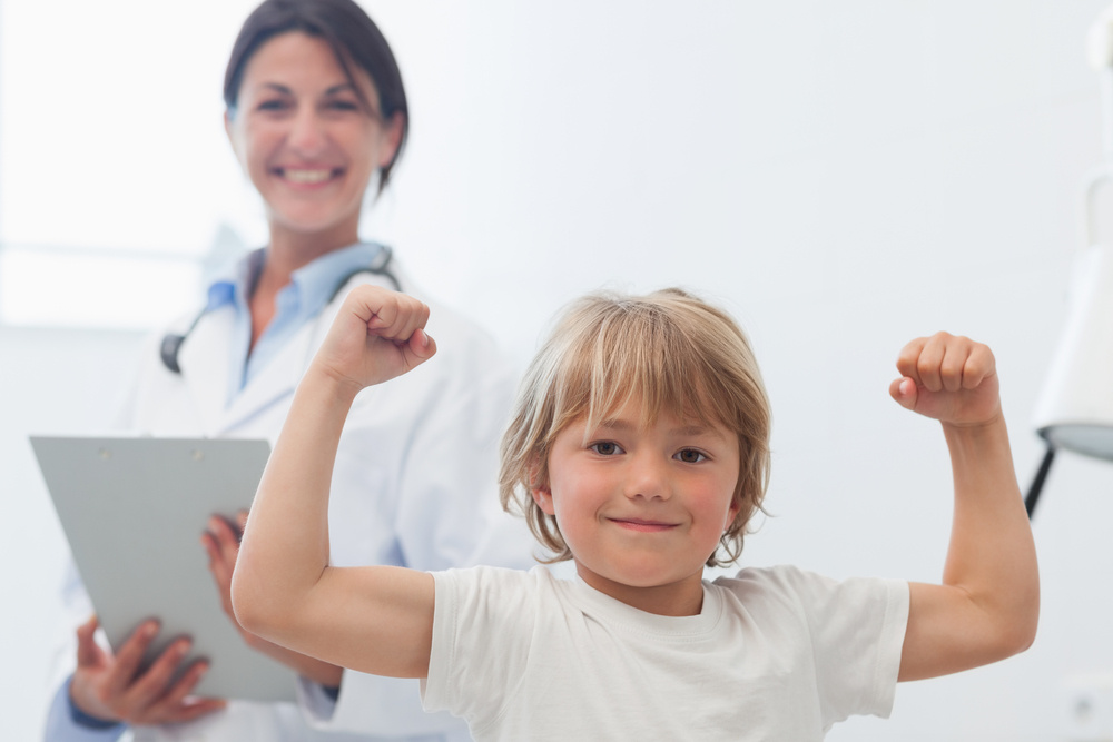 blonde child in white shirt showing off muscles with a female medical professional smiling and standing behind him