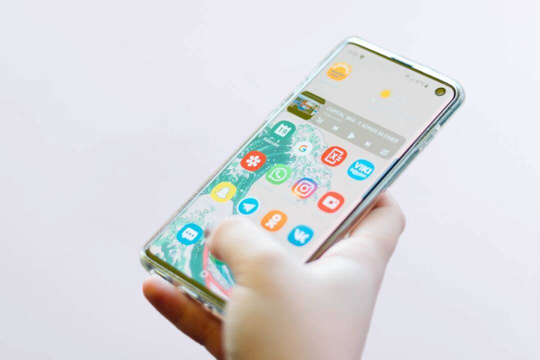 hand holding cell phone with home screen with apps visible and thumb on screen