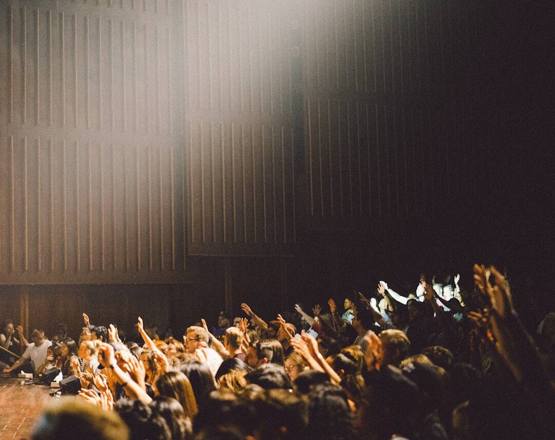 side of audience staring at stage with lights coming from stage in dark room and brown striped wall