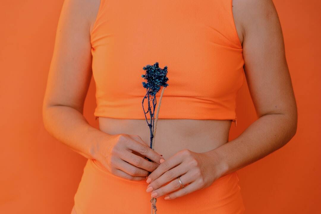 girl in orange shirt and pants with abdomen exposed holding a flower in front of her abdomen in front of an orange background