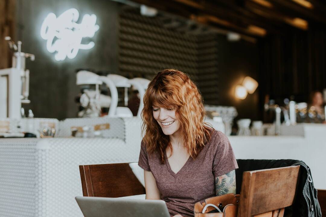 girl in front of laptop with reddish brown hair and light brown shirt in a blurry coffee shop background