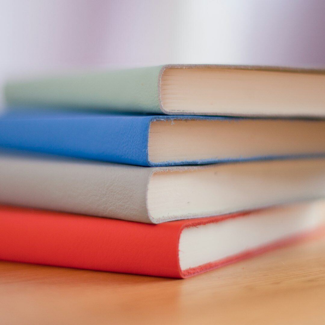 four books stacked on wooden desk from bottom to top colors of books orange grey blue and green 