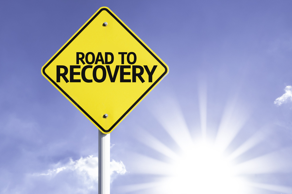 yellow roadsign that reads road to recovery with blue sky and sun in background